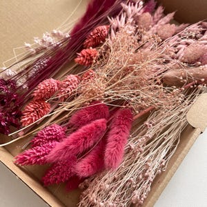 Op de afbeelding: Een kartonnen doos gevuld met gedroogde bloemen in roze en rode tinten. De opstelling bevat pluizige, veervormige en stekelige texturen. De bloemen zijn gerangschikt in verschillende vormen en maten.