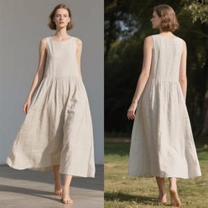 May include: A sleeveless, beige linen dress with a round neckline and a gathered waist. The dress falls to a midi length, creating a flowing silhouette. The fabric appears lightweight and breathable, suitable for warm weather.