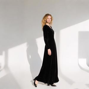May include: A long, black velvet dress with a V-neck and long sleeves. The dress has a flowing skirt and is worn with black heels. The model is standing in front of a white background.