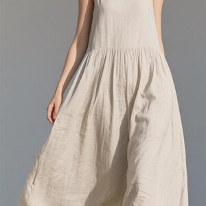 May include: A light beige linen dress with a sleeveless design and a gathered waist. The dress has a flowing, ankle-length skirt. The model is standing in front of a light gray background.