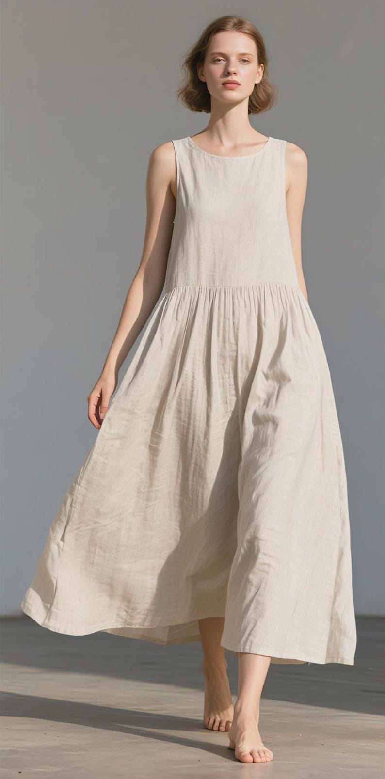 May include: A light beige linen dress with a sleeveless design and a gathered waist. The dress has a flowing, ankle-length skirt. The model is standing in front of a light gray background.