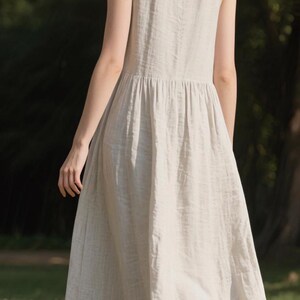 May include: A sleeveless, light beige linen dress with a gathered waist and a flowing skirt. The dress is knee-length and appears to be made of a lightweight fabric. The model is standing on grass in a natural outdoor setting.