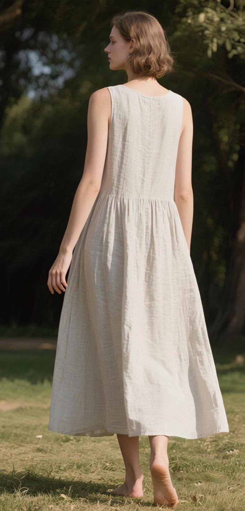May include: A sleeveless, light beige linen dress with a gathered waist and a flowing skirt. The dress is knee-length and appears to be made of a lightweight fabric. The model is standing on grass in a natural outdoor setting.