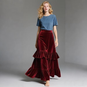 May include: A woman wearing a denim blue short-sleeved top and a long, tiered, burgundy velvet skirt. The skirt has a ruffled hem and falls to the floor. The outfit is set against a neutral gray background.