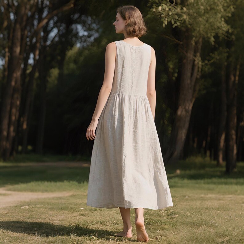May include: A sleeveless, light-colored dress with a gathered waist and a flowing skirt. The dress is made of a textured fabric and falls to mid-calf length. The person is walking on grass in a park-like setting.