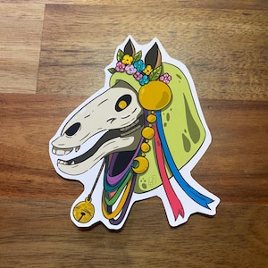 Op de afbeelding: Een sticker met een gestileerde paardenschedel met een bloemenkrans en een groene cape. De schedel is wit met zwarte oogkassen en een grijnzende mond. Kleurrijke linten en een gouden bel hangen aan de schedel.