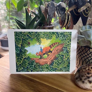 Puede incluir: Una ilustración en acuarela sobre una tarjeta blanca representa un panda rojo con una persona montada sobre su espalda, ambientada en un exuberante bosque verde. La obra de arte está enmarcada por follaje verde, con una superficie de madera en primer plano y objetos decorativos en el fondo.