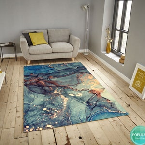 K&ouml;nnte beinhalten: Abstrakter Teppich mit einem blauen, goldenen und braunen Marmormuster. Der Teppich liegt auf einem Holzboden in einem Wohnzimmer.