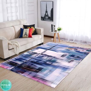May include: Abstract area rug with a geometric design in shades of blue, purple, pink, and white. The rug is laid out on a wooden floor in a living room setting.