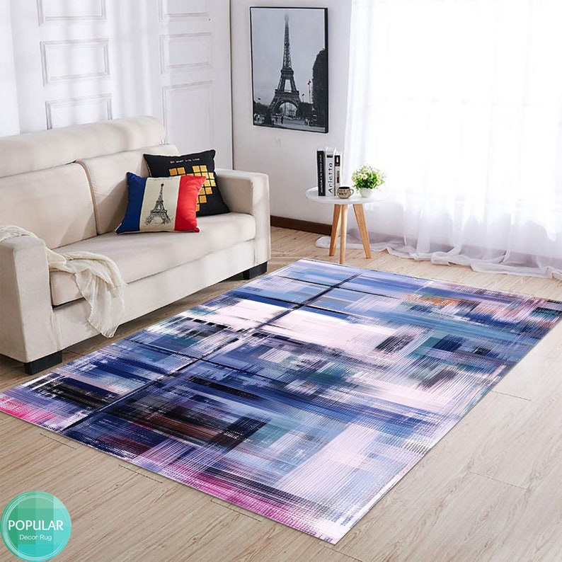 May include: Abstract area rug with a geometric design in shades of blue, purple, pink, and white. The rug is laid out on a wooden floor in a living room setting.