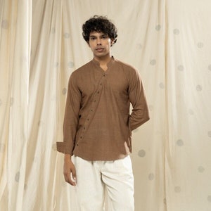 Puede incluir: Una camisa marrón de manga larga con cuello mandarín y cierre de botones en diagonal. La camisa se lleva con pantalones blancos y zapatos negros.