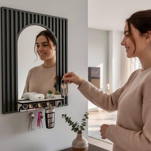 May include: A black framed wall mirror with an arched mirror and a shelf with hooks. The mirror has a black frame with vertical lines. The shelf holds small plants and keys. A person is reaching for the keys.
