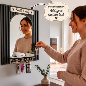 May include: A black wall-mounted mirror with vertical slats and a small shelf. The mirror features a light-coloured wooden plaque at the top with the text "Smith Family" and two heart icons. Below the shelf are four metal hooks for hanging keys.