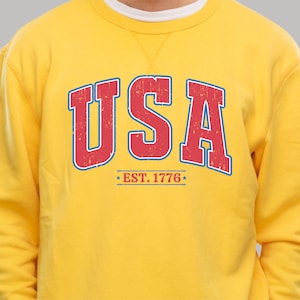 Op de afbeelding: Geel sweatshirt met een rode en blauwe vintage-stijl grafische afbeelding met de tekst "USA EST. 1776".