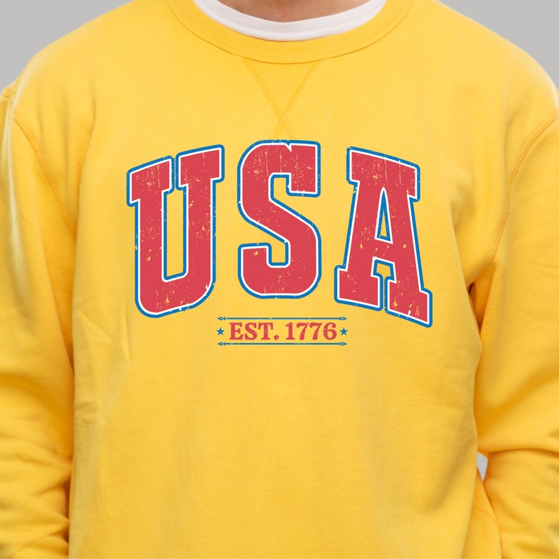 Op de afbeelding: Geel sweatshirt met een rode en blauwe vintage-stijl grafische afbeelding met de tekst "USA EST. 1776".