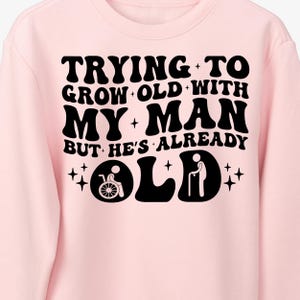 May include: Pink sweatshirt with black text that reads "Trying to grow old with my man but he's already old". The text is in a retro font style. There are two small black stick figures, one in a wheelchair and one with a cane, below the text.