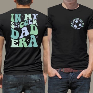 Op de afbeelding: Zwart t-shirt met de tekst "IN MY SOCCER DAD ERA" in retrostijl, met een voetbalafbeelding. Het ontwerp gebruikt blauwe en groene tinten. Het t-shirt wordt door een persoon gedragen.