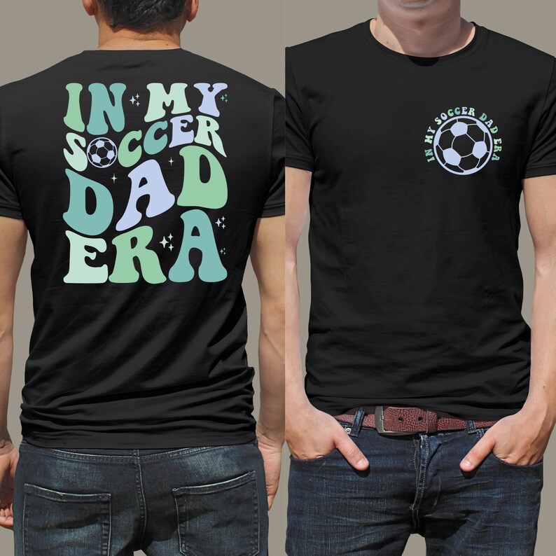 Op de afbeelding: Zwart t-shirt met de tekst "IN MY SOCCER DAD ERA" in retrostijl, met een voetbalafbeelding. Het ontwerp gebruikt blauwe en groene tinten. Het t-shirt wordt door een persoon gedragen.