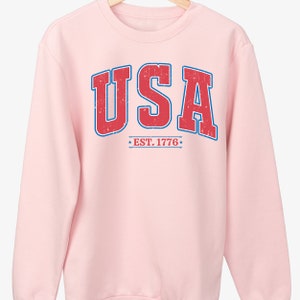 Op de afbeelding: Roze sweatshirt met een vintage-stijl "USA EST. 1776" grafiek in rood en blauw.