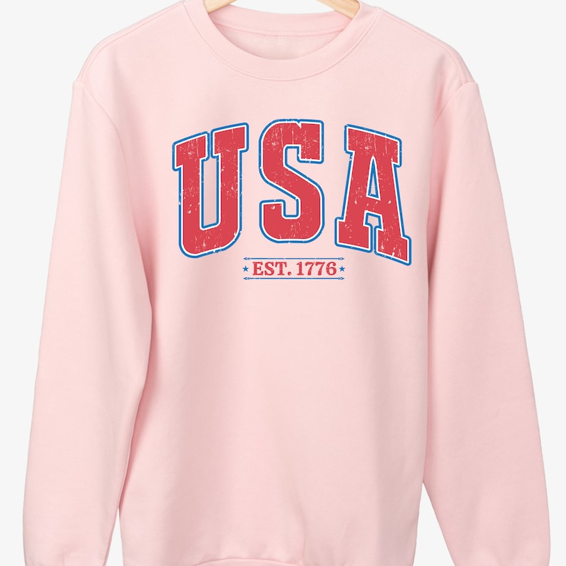 Op de afbeelding: Roze sweatshirt met een vintage-stijl "USA EST. 1776" grafiek in rood en blauw.