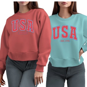 Op de afbeelding: Twee vrouwen dragen sweatshirts met het woord "USA" in rode en blauwe letters. Het sweatshirt aan de linkerkant is koraal en het sweatshirt aan de rechterkant is lichtblauw. Het sweatshirt aan de rechterkant heeft de tekst "EST. 1776" onder het woord "USA".
