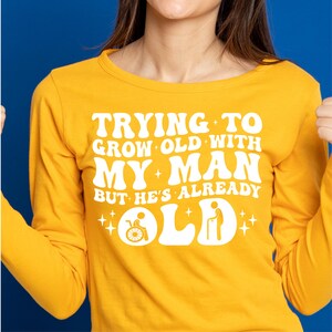 May include: A yellow long-sleeved shirt with white text that reads "Trying to grow old with my man but he's already old". The text is in a retro font style with a graphic of a person in a wheelchair and a person with a cane.