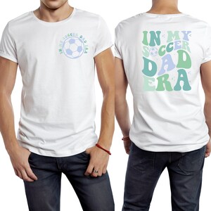 Op de afbeelding: Wit T-shirt met opgerolde mouwen. De voorkant heeft een voetbalafbeelding met de tekst "IN MY SOCCER DAD ERA" in blauw en groen. De achterkant van de shirt heeft dezelfde tekst in een groter, retro lettertype. Het shirt wordt gecombineerd met een blauwe jeans.