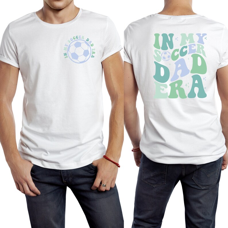 Op de afbeelding: Wit T-shirt met opgerolde mouwen. De voorkant heeft een voetbalafbeelding met de tekst "IN MY SOCCER DAD ERA" in blauw en groen. De achterkant van de shirt heeft dezelfde tekst in een groter, retro lettertype. Het shirt wordt gecombineerd met een blauwe jeans.