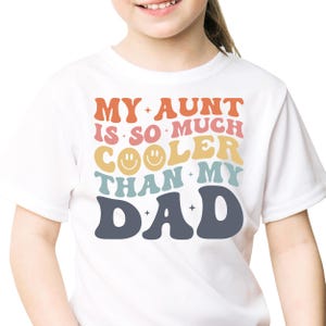 May include: White t-shirt with the text "MY AUNT IS SO MUCH COOLER THAN MY DAD" in orange, pink, yellow, teal, and navy blue. The word "COOLER" has two smiley faces. The shirt is for children.