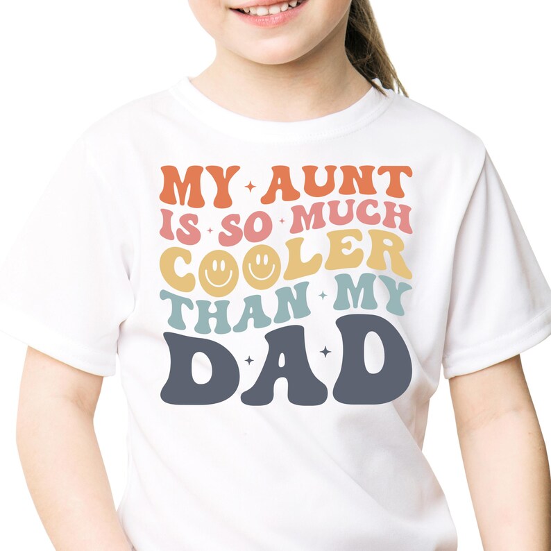 May include: White t-shirt with the text "MY AUNT IS SO MUCH COOLER THAN MY DAD" in orange, pink, yellow, teal, and navy blue. The word "COOLER" has two smiley faces. The shirt is for children.