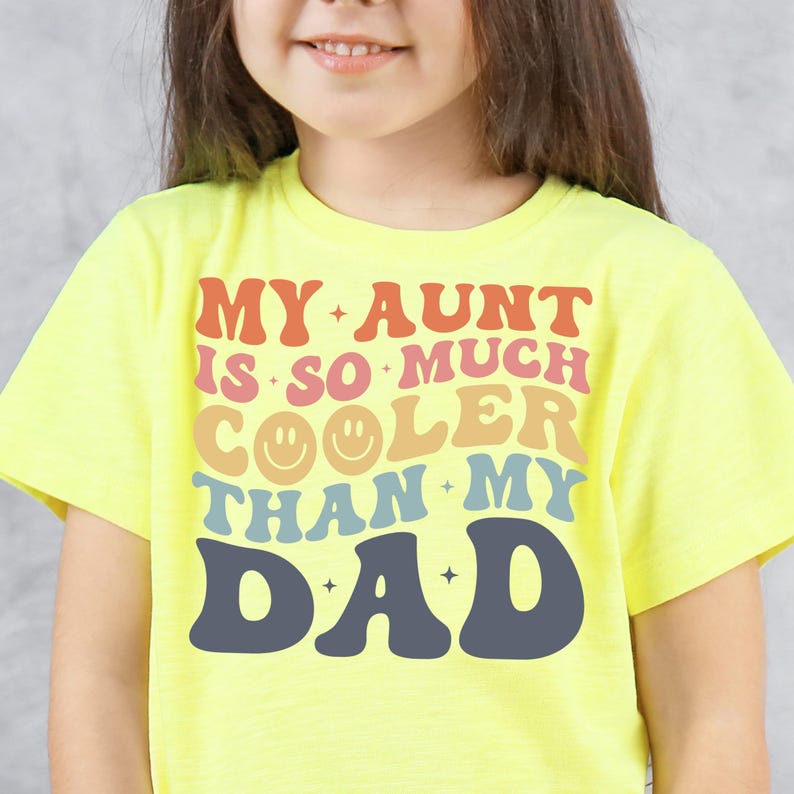 May include: A yellow t-shirt with the text "MY AUNT IS SO MUCH COOLER THAN MY DAD" in retro-style lettering. The design includes a smiley face and star accents. The shirt is a bright, cheerful color.