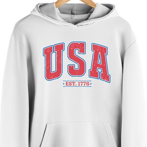 Op de afbeelding: Witte hoodie met een rode en blauwe vintage-stijl grafische afbeelding met de tekst "USA EST. 1776".