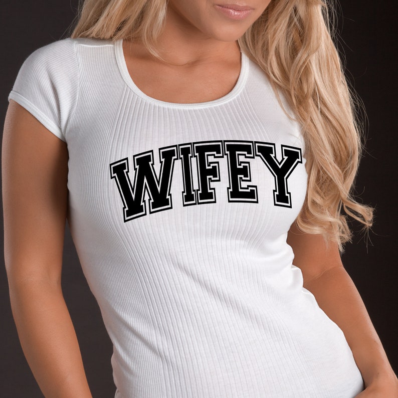 May include: White ribbed t-shirt with the word "WIFEY" in bold black letters. The shirt has a scoop neck and short sleeves. The text is in a collegiate style font.
