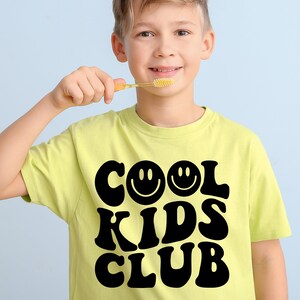 Peut inclure: Un jeune portant un t-shirt jaune avec le texte "Cool Kids Club" en lettres noires. Deux visages souriants font partie du texte. La personne tient une brosse &agrave; dents.