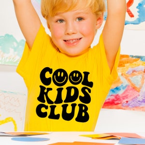Peut inclure: Un jeune enfant portant un t-shirt jaune avec le texte "COOL KIDS CLUB" en lettres noires avec deux visages souriants. Le t-shirt a une police de caract&egrave;res r&eacute;tro des ann&eacute;es 70.