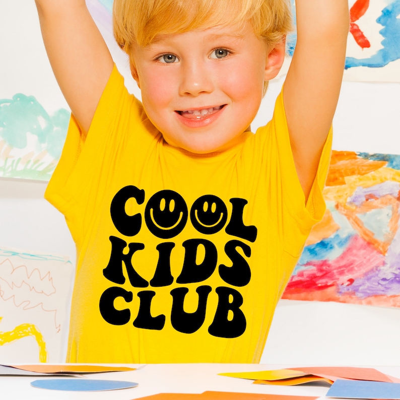 Peut inclure: Un jeune enfant portant un t-shirt jaune avec le texte "COOL KIDS CLUB" en lettres noires avec deux visages souriants. Le t-shirt a une police de caract&egrave;res r&eacute;tro des ann&eacute;es 70.