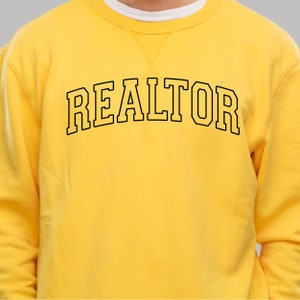 Peut inclure: Sweat-shirt jaune avec un texte noir qui dit "REALTOR" dans une police d'&eacute;criture universitaire.