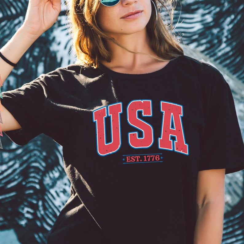 Op de afbeelding: Een zwart T-shirt met een rode en blauwe "USA"-afbeelding en de tekst "EST. 1776".