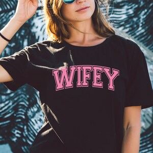 May include: Black crew neck t-shirt with the word "WIFEY" in pink collegiate-style lettering. The text is outlined in a darker pink. The shirt is worn by a person with sunglasses and a black bracelet.