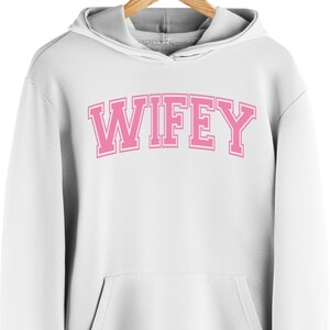 May include: White hooded sweatshirt with the word "WIFEY" in pink block letters across the chest. The letters have a white outline. The sweatshirt has a front pocket and a gray-lined hood.