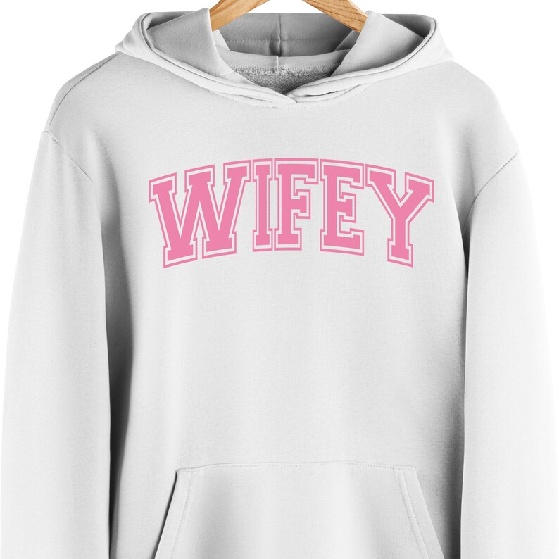 May include: White hooded sweatshirt with the word "WIFEY" in pink block letters across the chest. The letters have a white outline. The sweatshirt has a front pocket and a gray-lined hood.