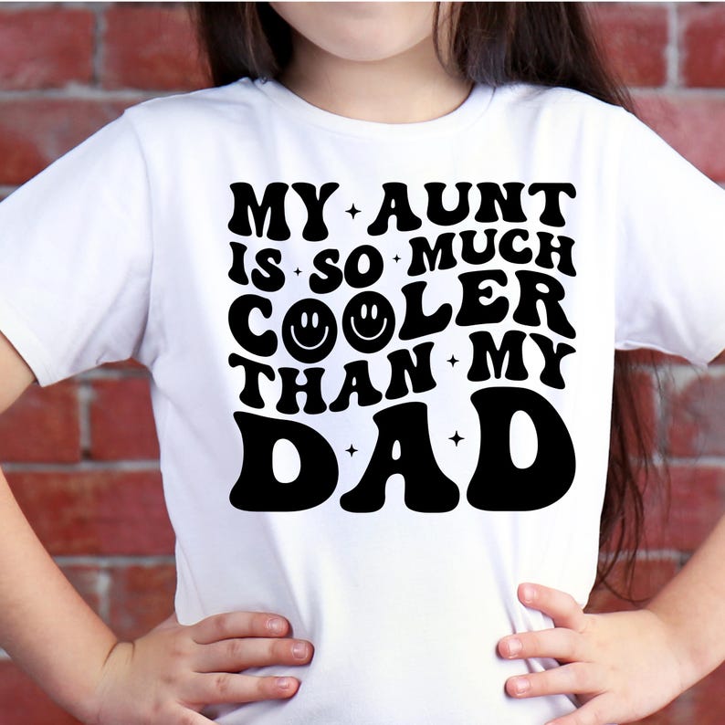 May include: White t-shirt with the text "MY AUNT IS SO MUCH COOLER THAN MY DAD" in black, bold, retro-style font. The word "COOLER" features a smiley face. The shirt is worn by a person with their hands on their hips.