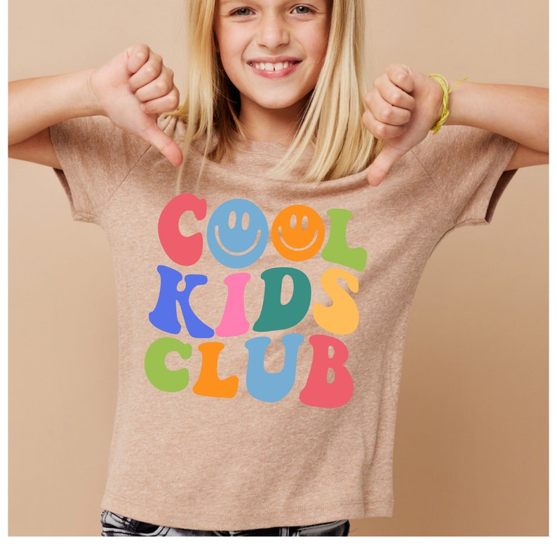 Peut inclure: Un t-shirt marron clair avec un imprim&eacute; graphique des mots "COOL KIDS CLUB" en lettres color&eacute;es avec des smileys.