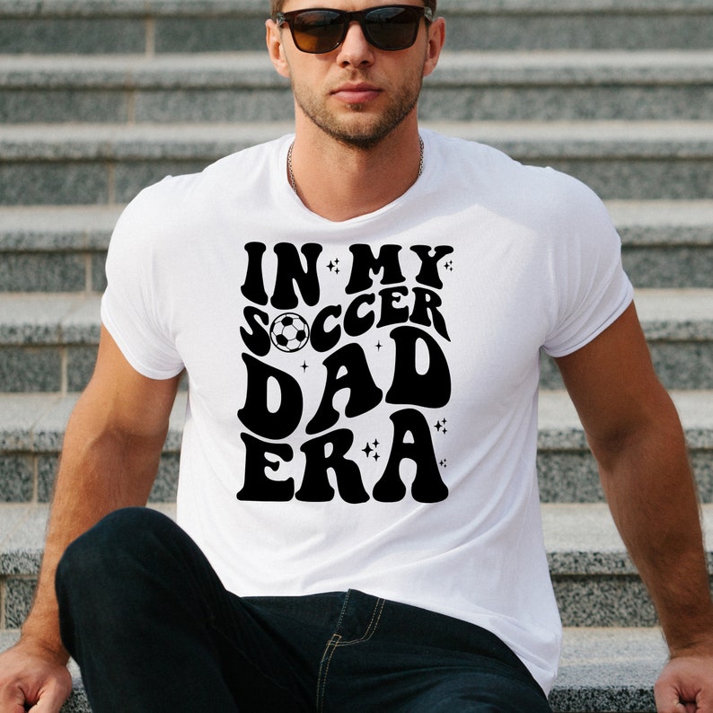 Op de afbeelding: Wit t-shirt met de zwarte tekst "IN MY SOCCER DAD ERA" in retro-stijl. Het ontwerp bevat een voetbal en sterren. Het t-shirt heeft een ronde hals.