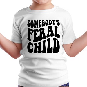 Może przedstawiać: Biała koszulka z czarnym napisem "Somebody's Feral Child" w retro czcionce.