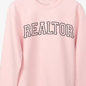 Peut inclure: Sweat-shirt rose avec le mot "REALTOR" imprim&eacute; en noir dans une police d'&eacute;criture universitaire.
