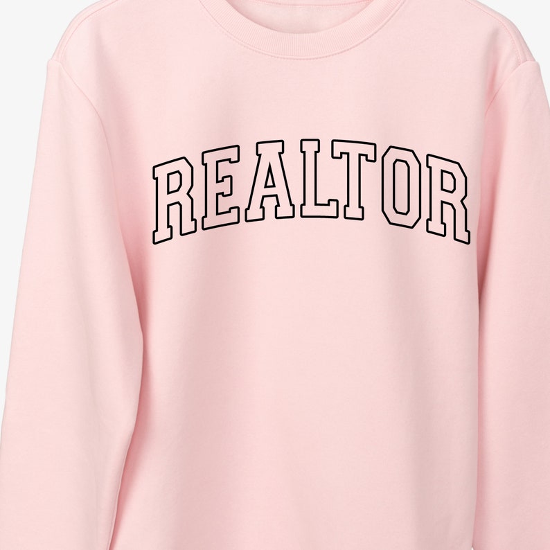 Peut inclure: Sweat-shirt rose avec le mot "REALTOR" imprim&eacute; en noir dans une police d'&eacute;criture universitaire.