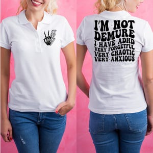 May include: White polo shirt with a black skeleton hand with a peace sign and lightning bolt on the left chest. The back of the shirt has black text that reads "I'm not demure I have ADHD very forgetful very chaotic very anxious".
