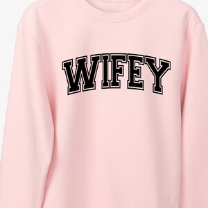 May include: A light pink sweatshirt with the word "WIFEY" printed across the chest in bold, black, collegiate-style lettering with a white outline. The sweatshirt has a crew neck and long sleeves.