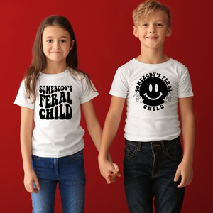 Może przedstawiać: Dwoje dzieci w białych koszulkach z czarnym nadrukiem. Koszulka dziewczynki ma napis "Somebody's Feral Child". Koszulka chłopca ma napis "Somebody's Feral Child" z uśmiechniętą buźką i znakami pokoju. Trzymają się za ręce i stoją przed czerwonym tłem.